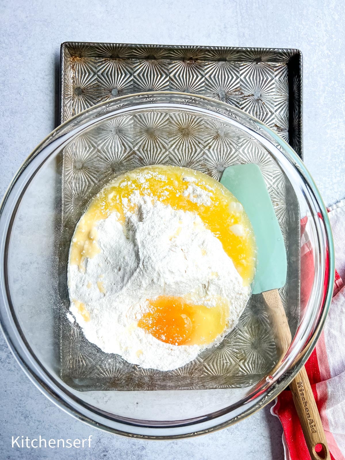 A glass bowl with flour, egg, and melted butter next to a spatula, on a patterned baking tray.
