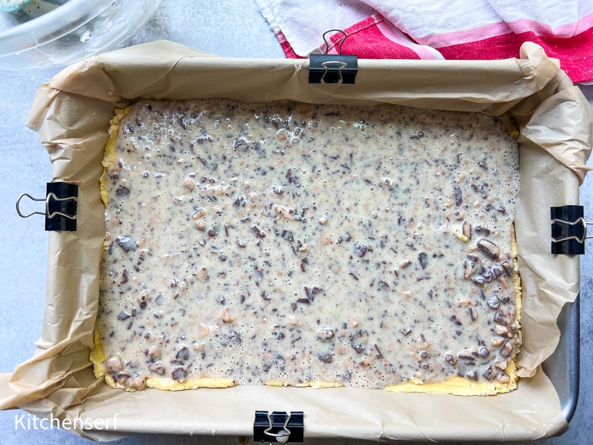 Unbaked dessert bars with a nutty mixture in a parchment-lined pan, secured with binder clips.