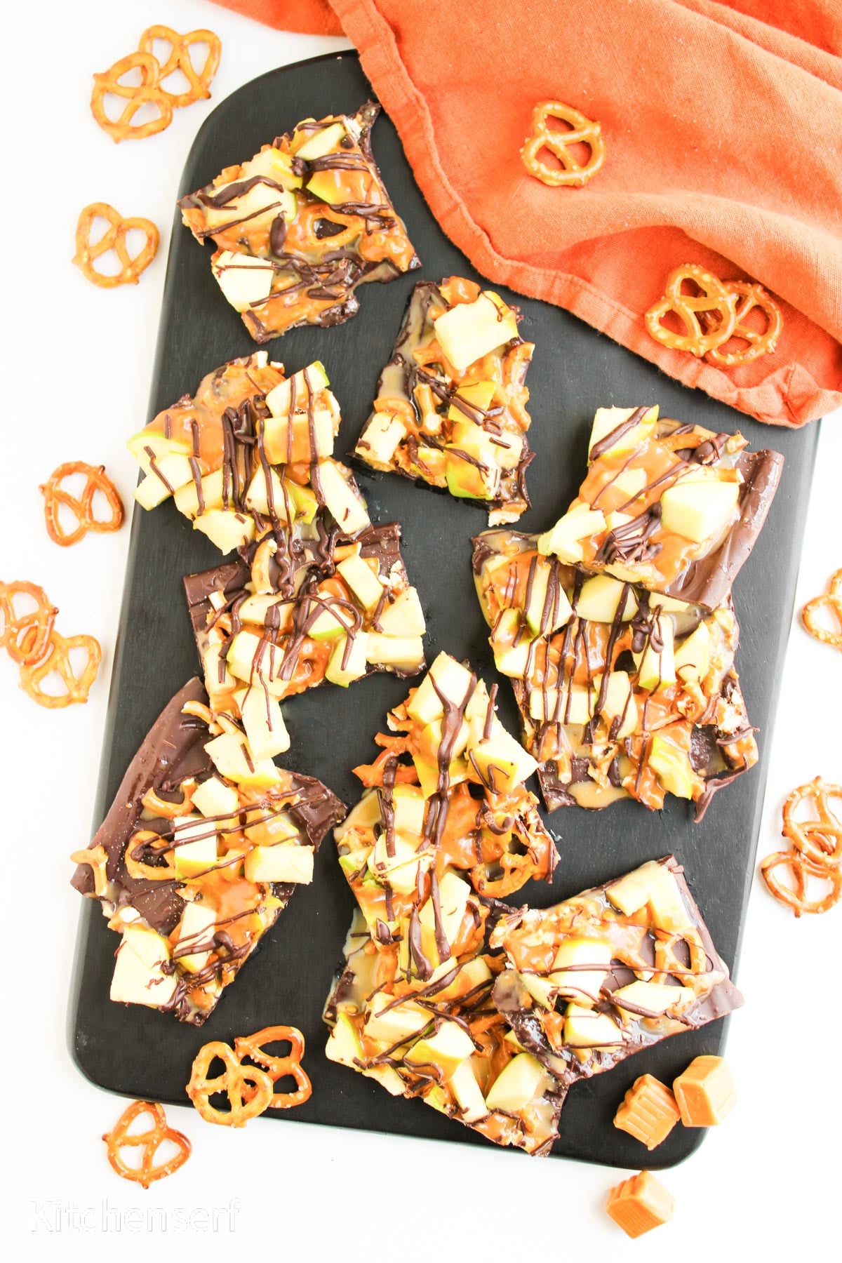 Chocolate bark topped with apple pieces, caramel, and pretzels on a black tray, with pretzels scattered around.