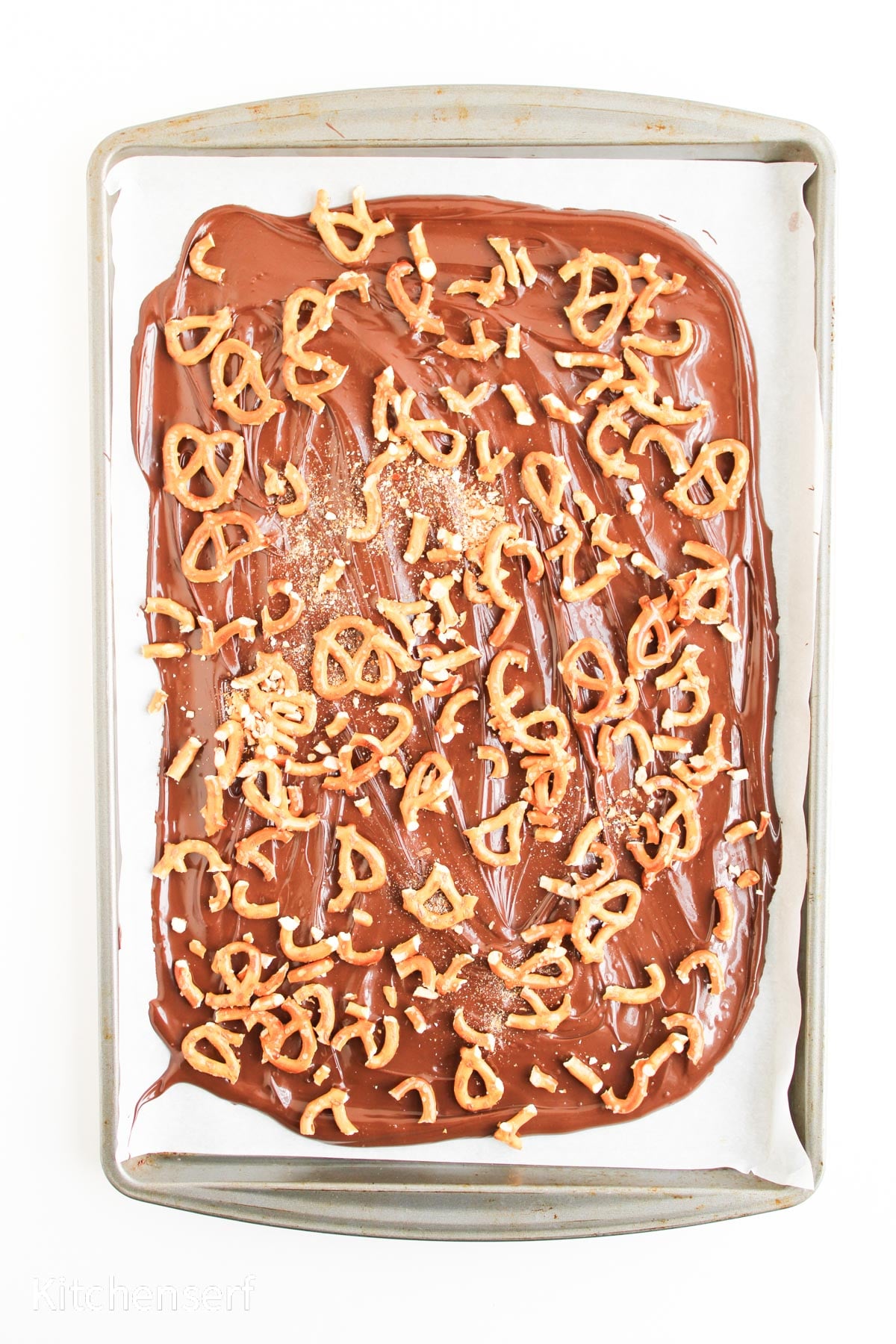 A sheet pan with melted chocolate topped with broken pretzel pieces on parchment paper.