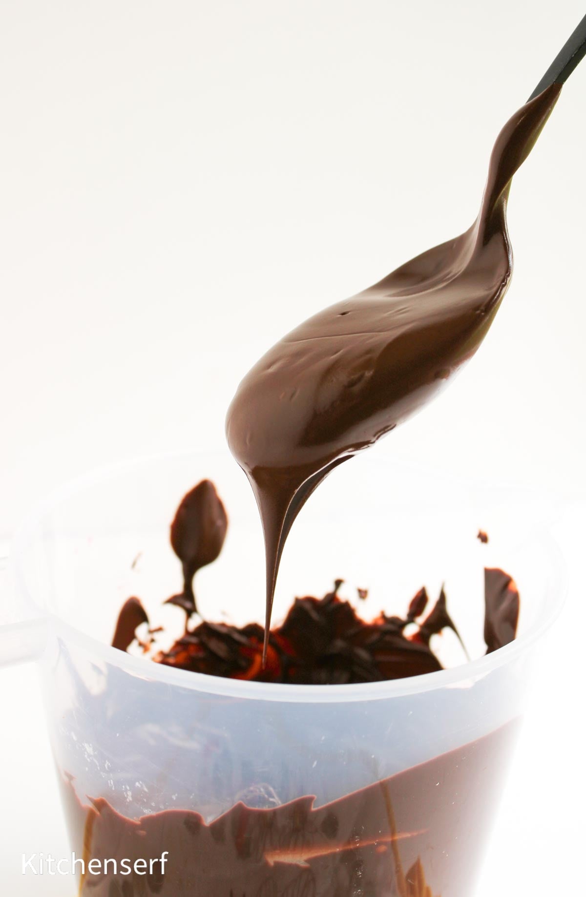 A spoon drips melted chocolate into a clear container against a white background.