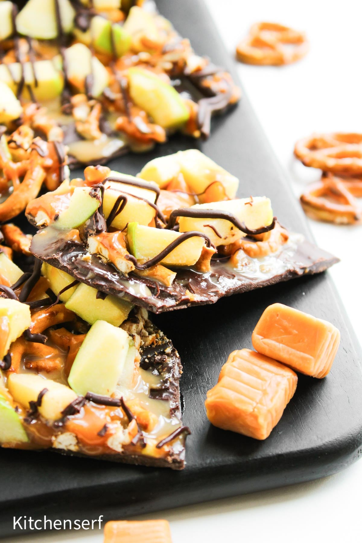 Chocolate bark topped with green apple pieces, pretzels, caramel, and a drizzle of chocolate on a black tray.