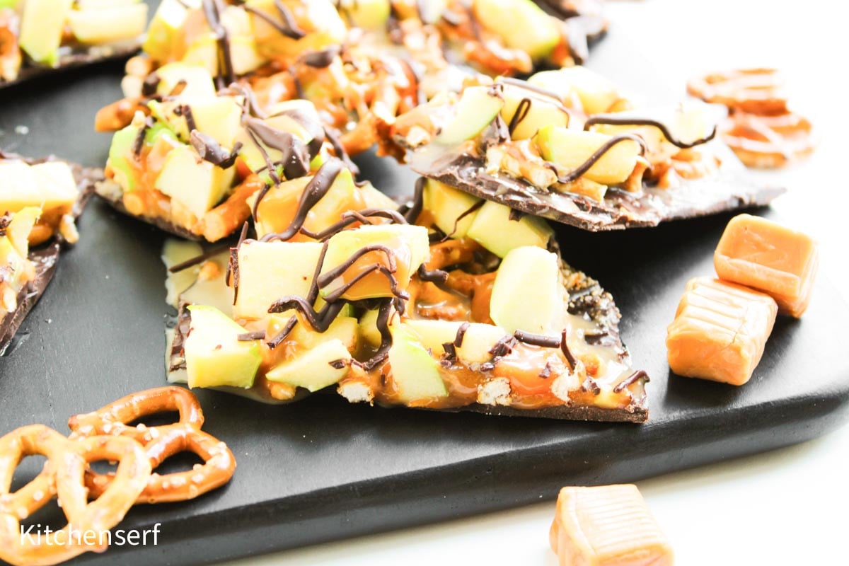 Chocolate bark topped with pretzels, caramel, apple pieces, and chocolate drizzle on a black tray.