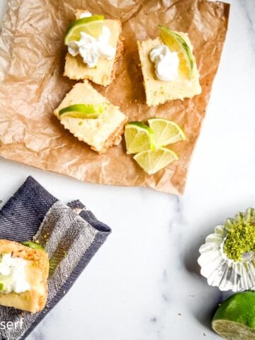 Top-down view of key lime pie bars topped with whipped cream and lime slices, arranged on parchment and a napkin, with fresh limes nearby.