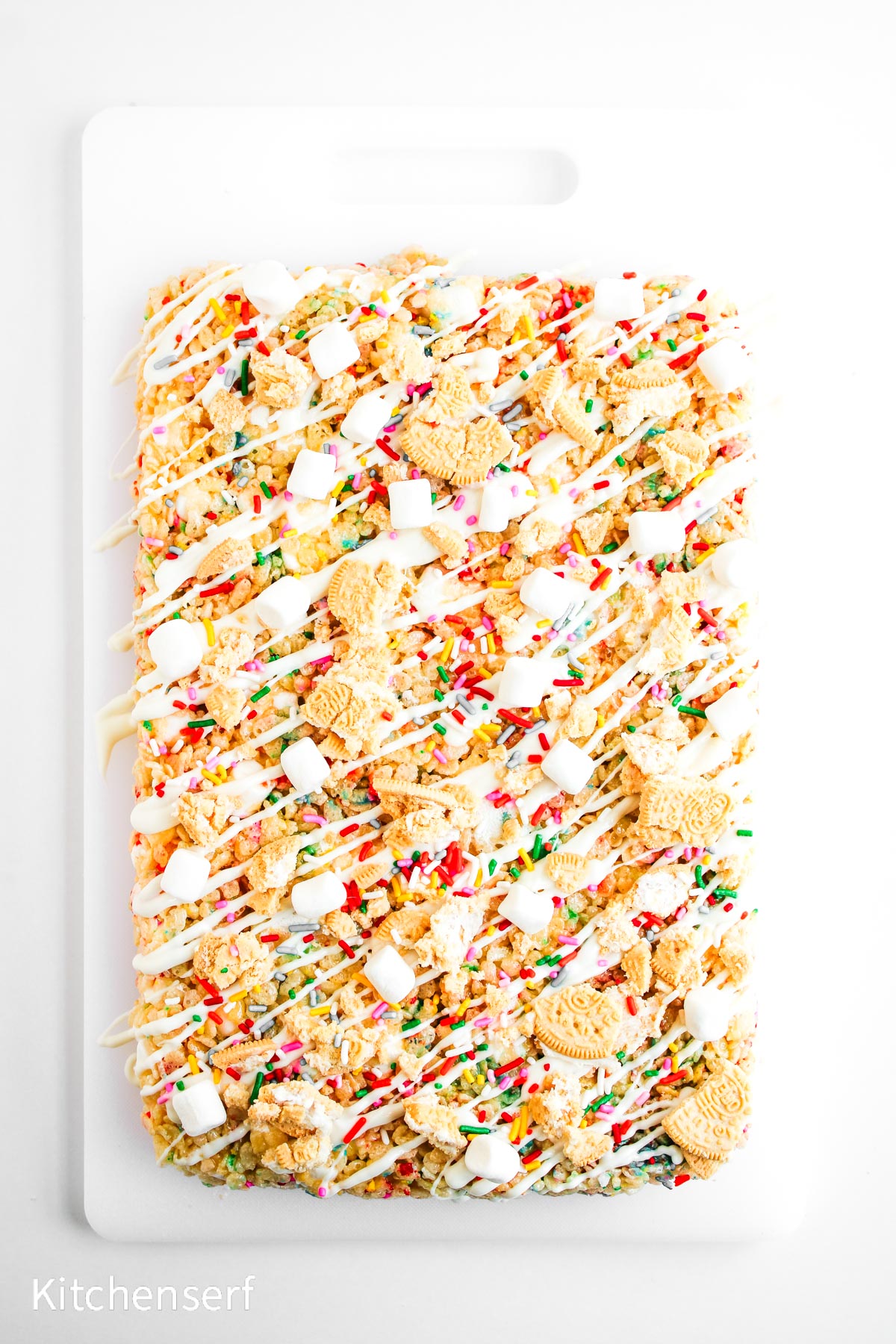 A rectangular dessert with cookies, marshmallows, sprinkles, and white drizzle on a white cutting board.