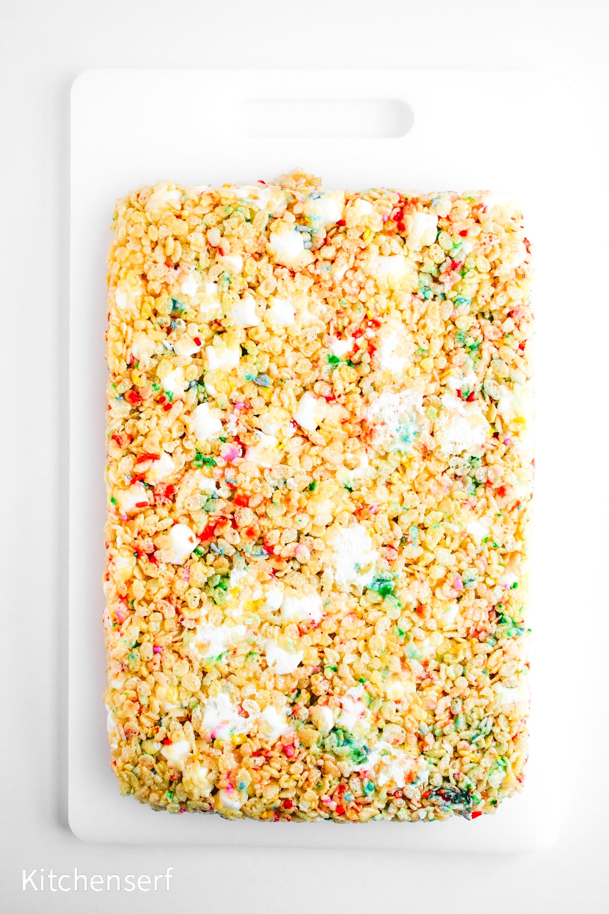 Rectangular rice crispy treat with colorful sprinkles and melted marshmallows on a white cutting board.