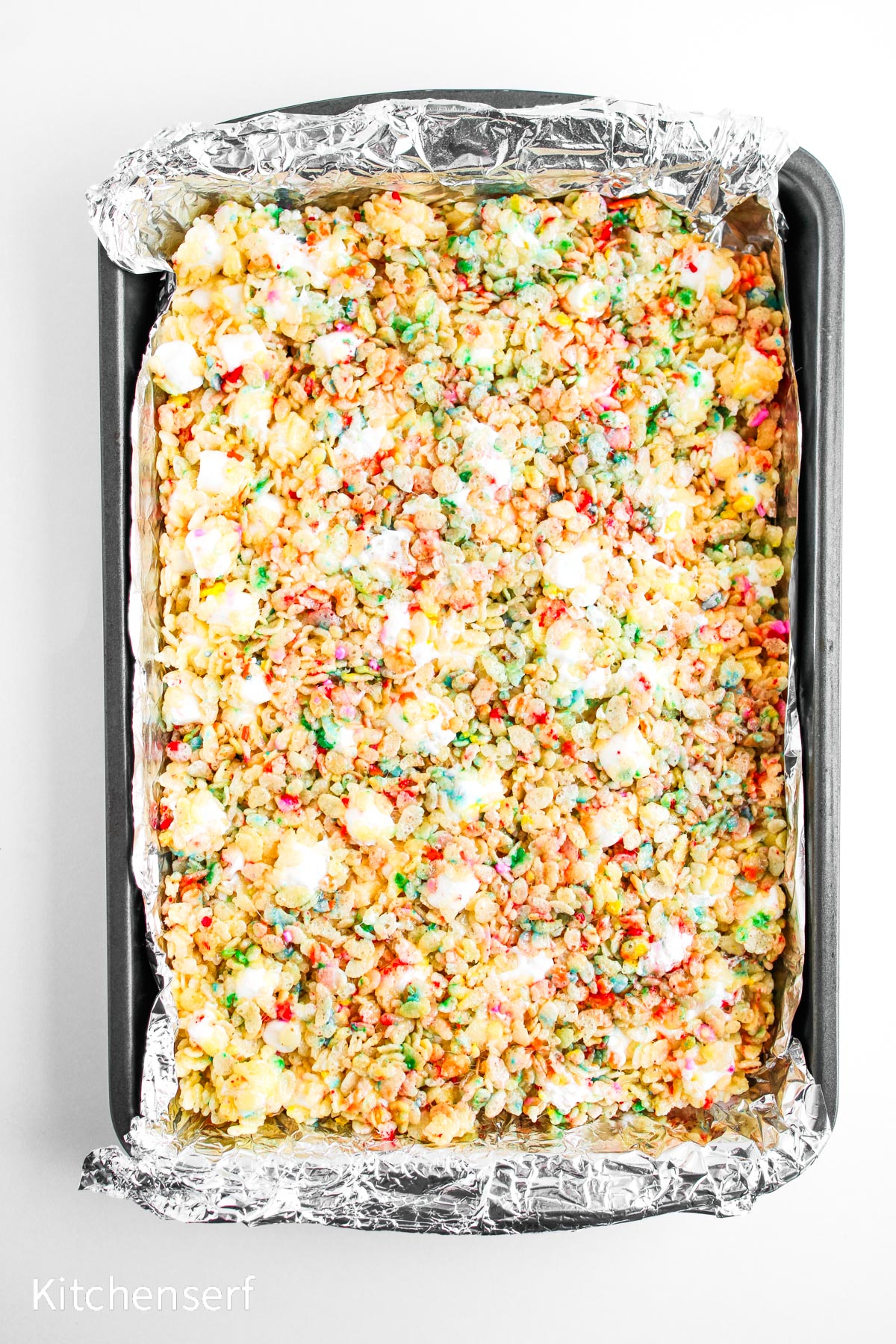 A foil-lined pan filled with colorful marshmallow rice cereal treats on a white background.