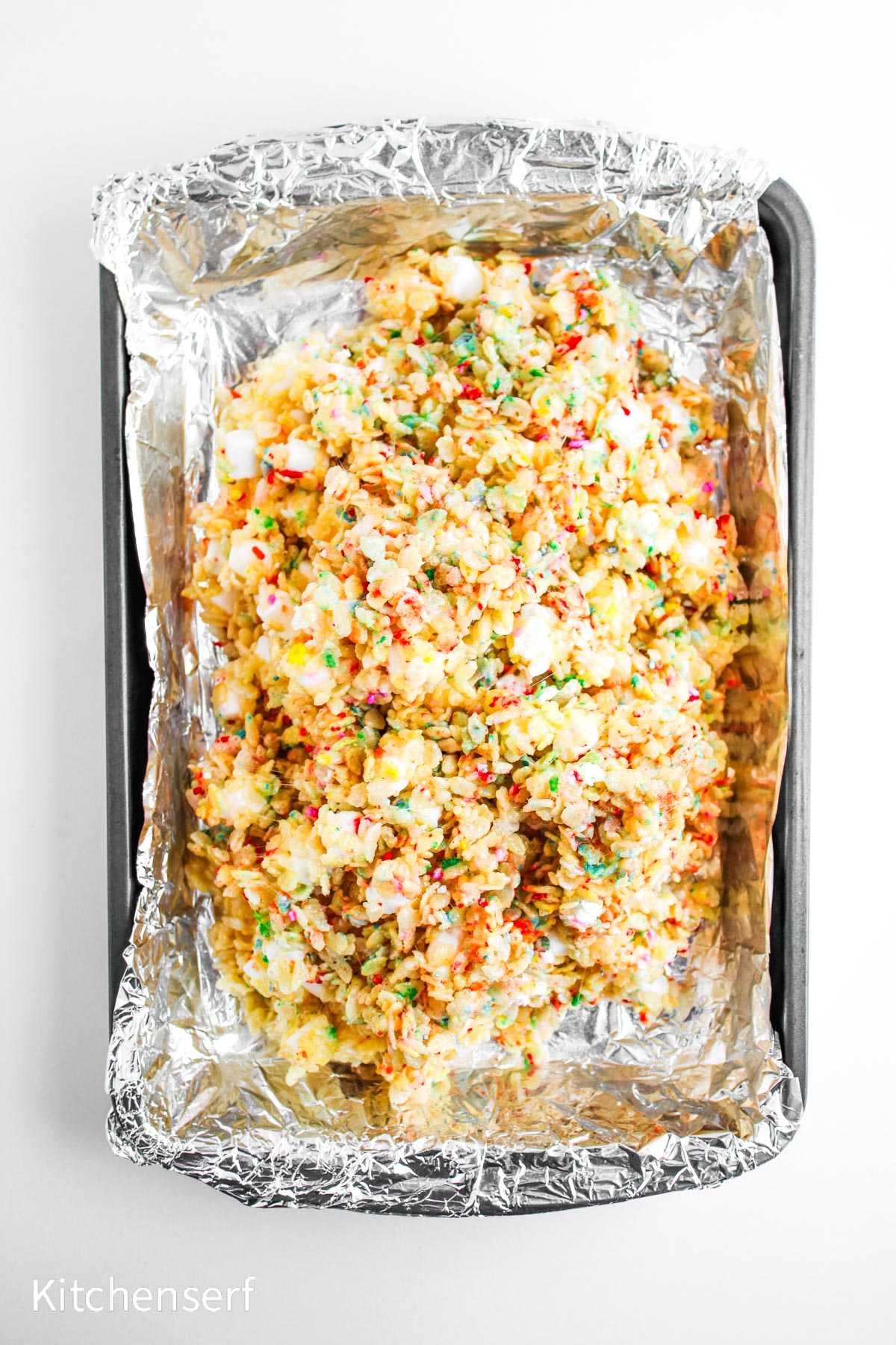 Colorful cereal marshmallow treats in a foil-lined baking pan on a white surface.