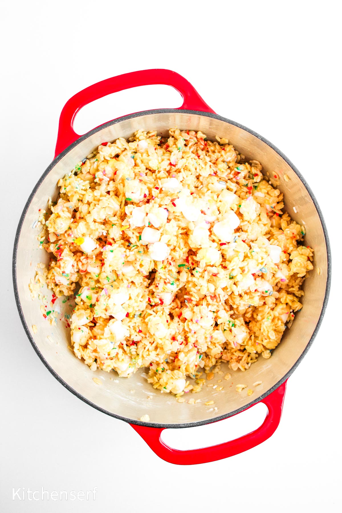 A red pot filled with rice krispie treats mixed with mini marshmallows on a white background.