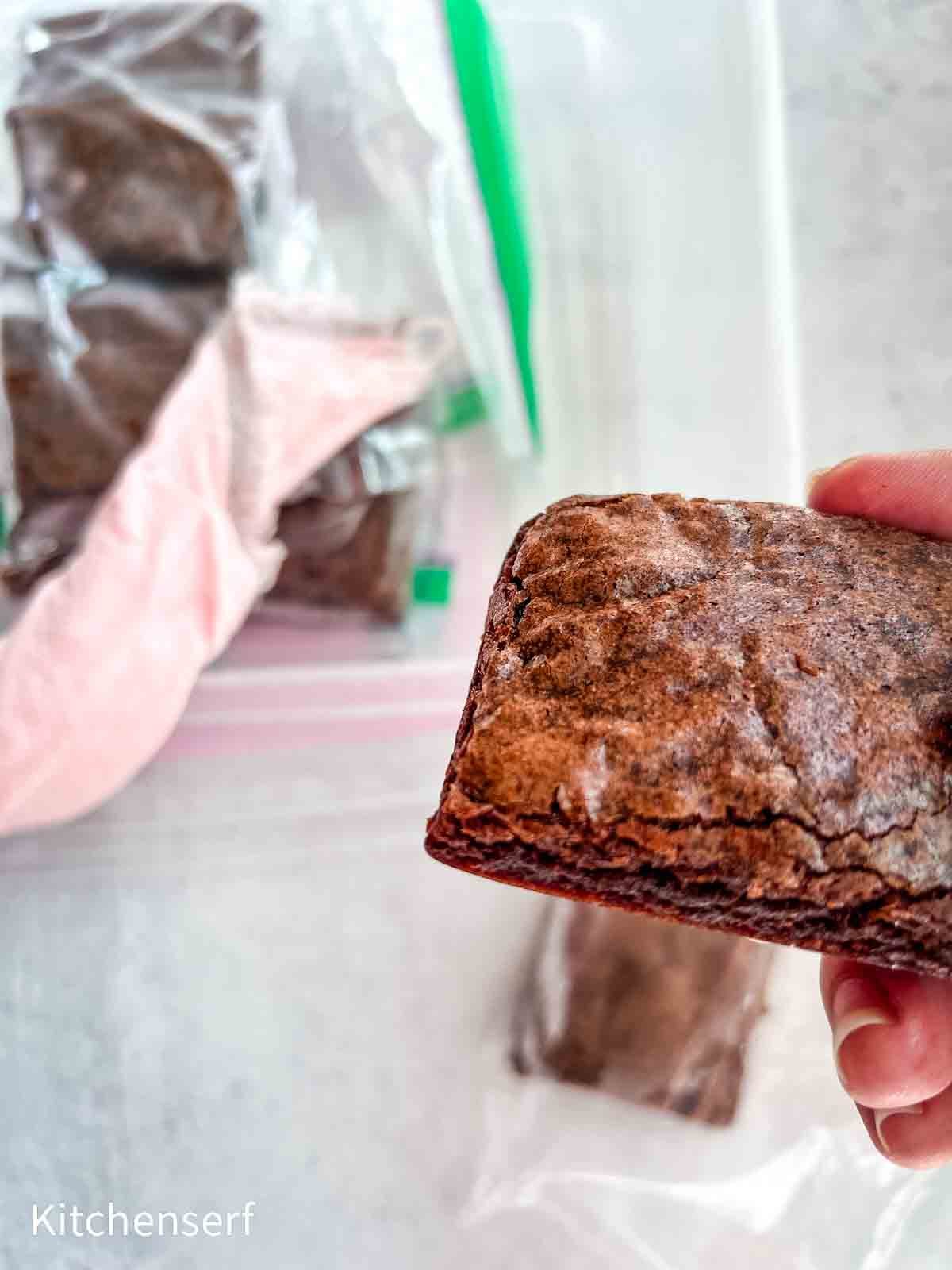 A hand holding a brownie with more brownies in ziplock bags on a white surface.