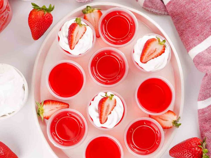 Red gelatin shots topped with whipped cream and strawberry slices on a white plate, surrounded by fresh strawberries.
