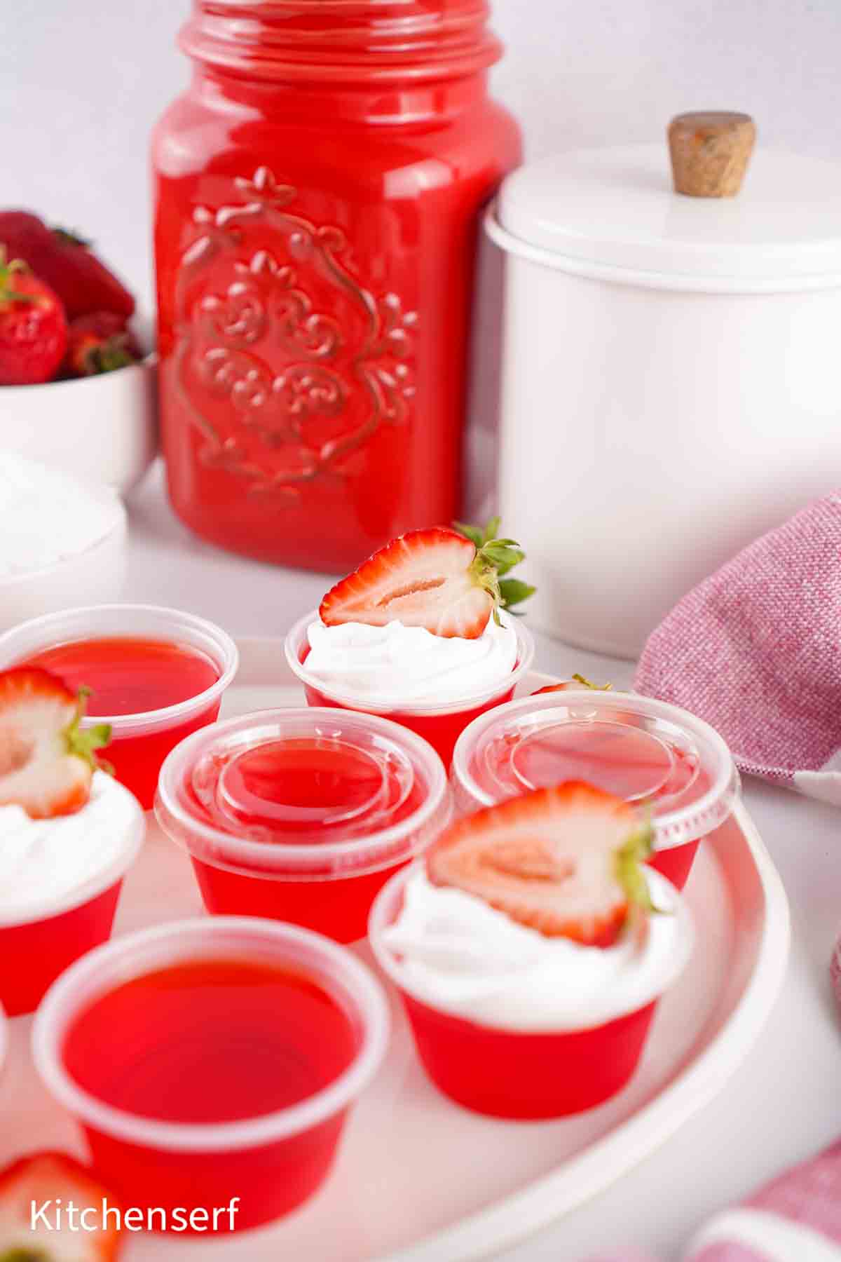 Small red gelatin cups topped with whipped cream and sliced strawberries, arranged on a white tray.