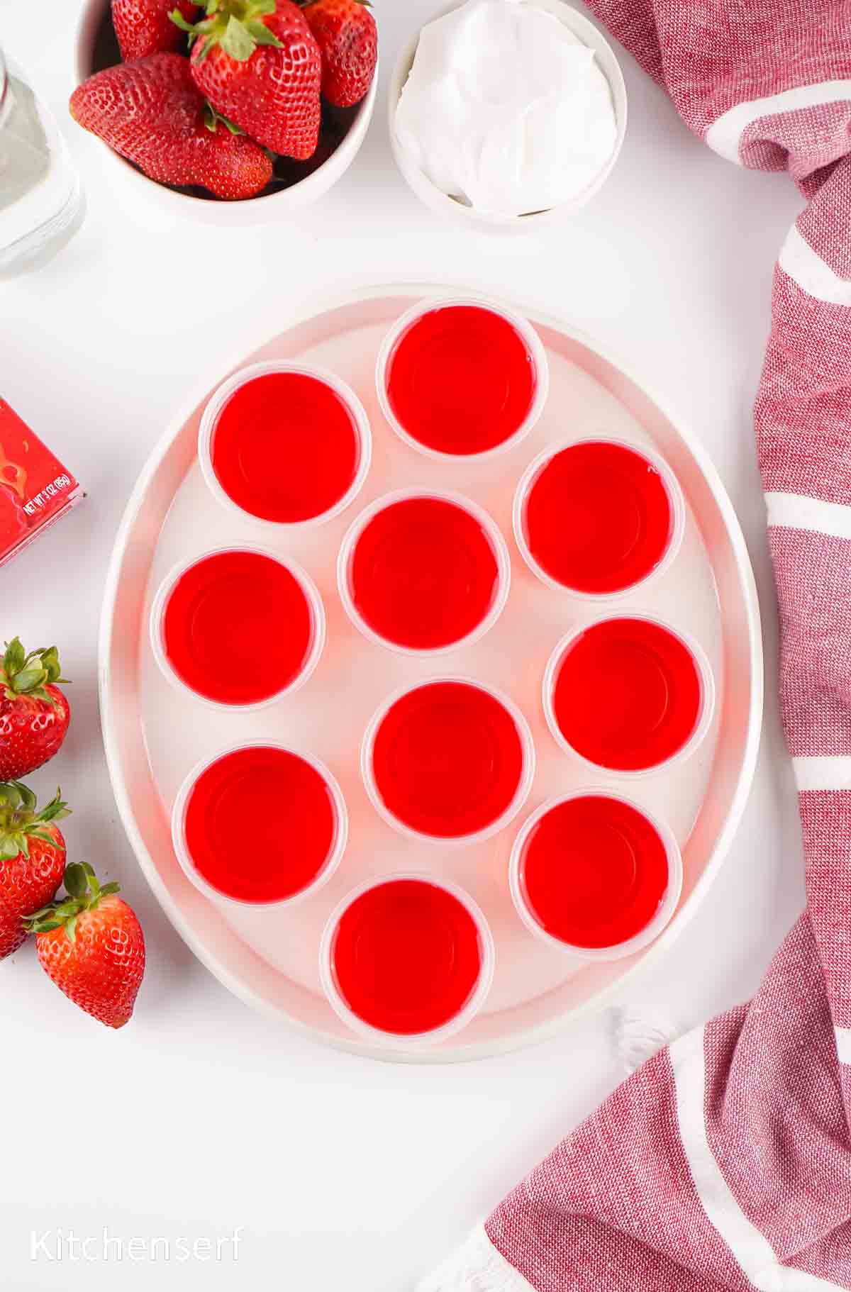 A tray of red gelatin shots in small cups, surrounded by strawberries, a towel, and a bowl of whipped cream.