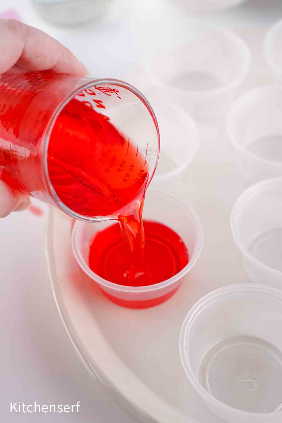 Red liquid being poured from a measuring cup into a small plastic cup, with empty cups nearby.