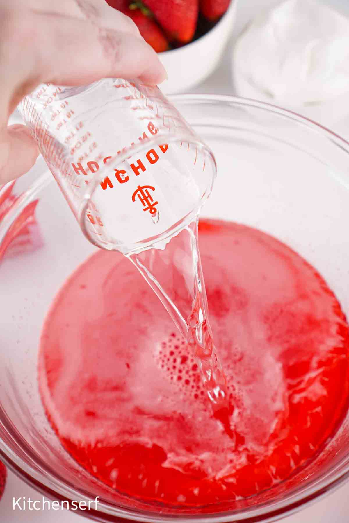 A hand pours clear liquid from a measuring cup into a bowl of red mixture.