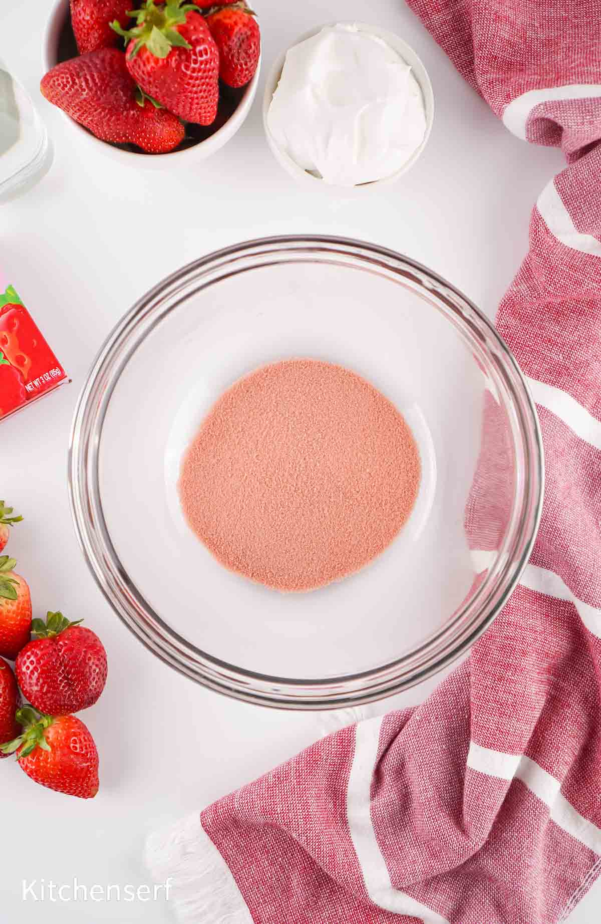 A glass bowl with pink powder, surrounded by strawberries, whipped topping, and a red-striped towel.