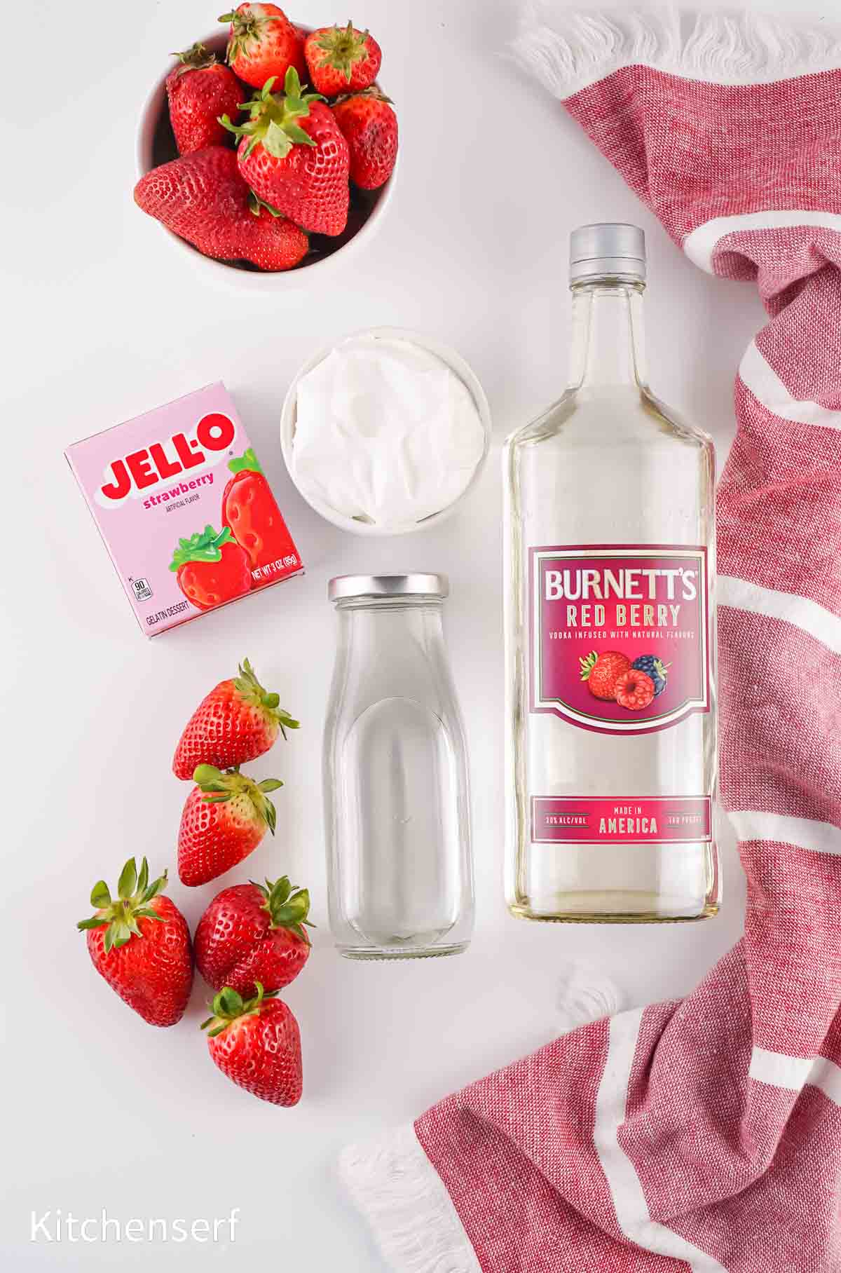 Strawberries, strawberry Jell-O, whipped topping, vodka, and milk on a white surface with a red striped towel.