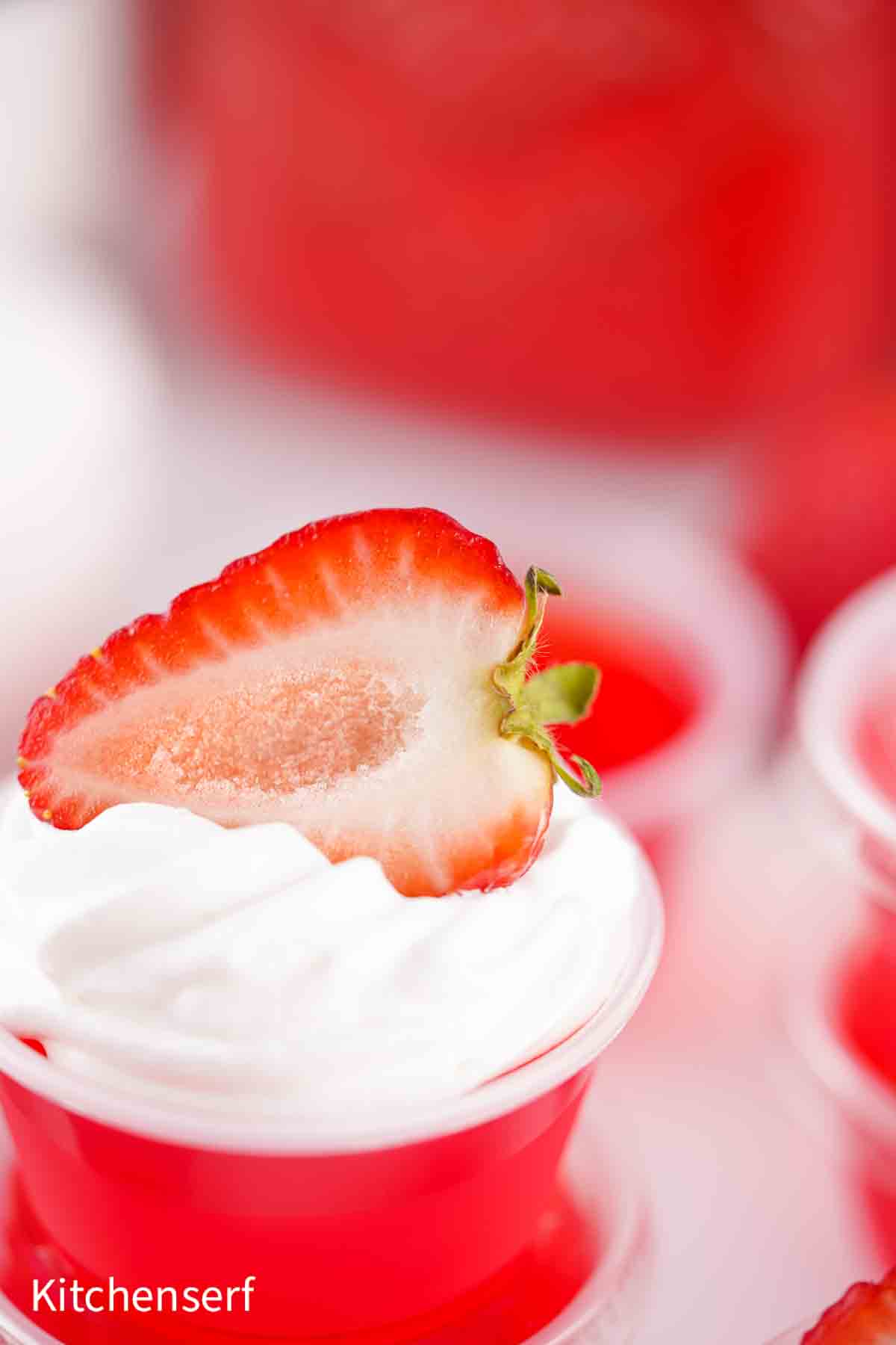 A strawberry half sits atop whipped cream on a red gelatin dessert cup.