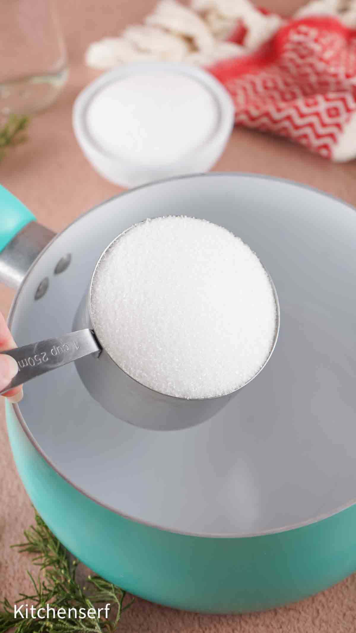 A metal measuring cup filled with sugar held over a teal saucepan on a kitchen counter.