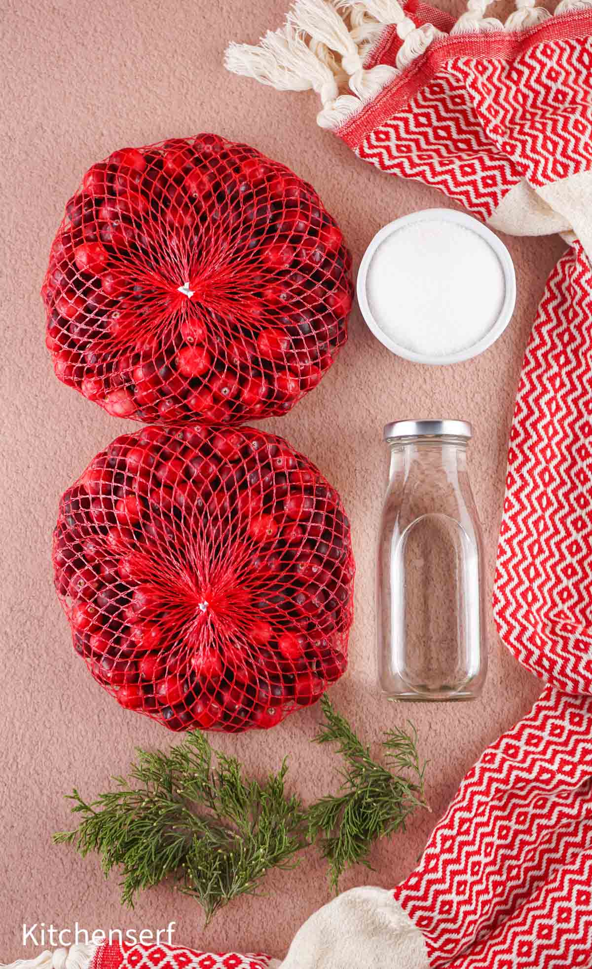 Two bags of cranberries, a bottle, a bowl of sugar, greenery, and red-patterned towels on a tan surface.