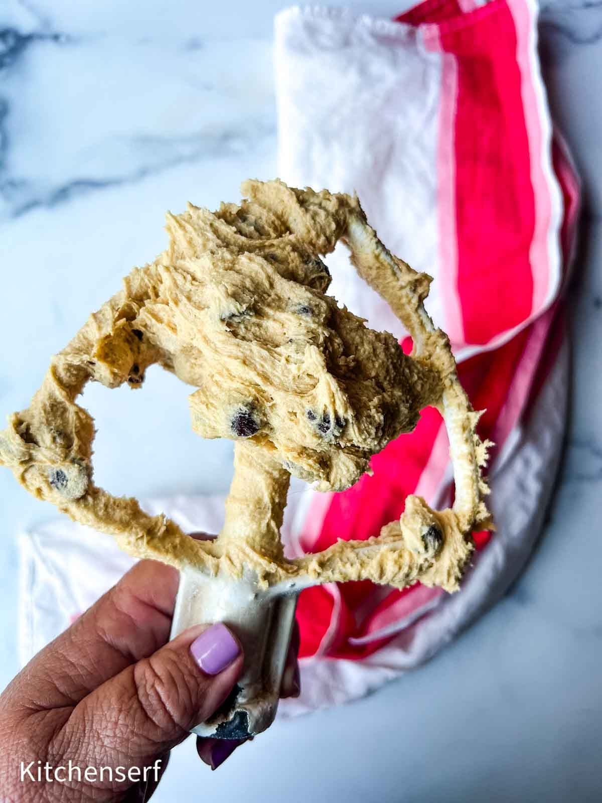 A hand holding a mixer beater covered in chocolate chip cookie dough above a red and white towel.