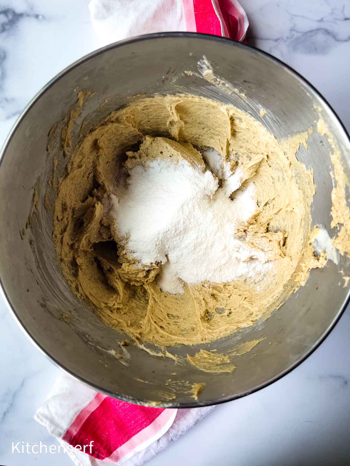 A metal mixing bowl with creamed batter and sugar on top, sitting on a marble counter with a red-striped towel.