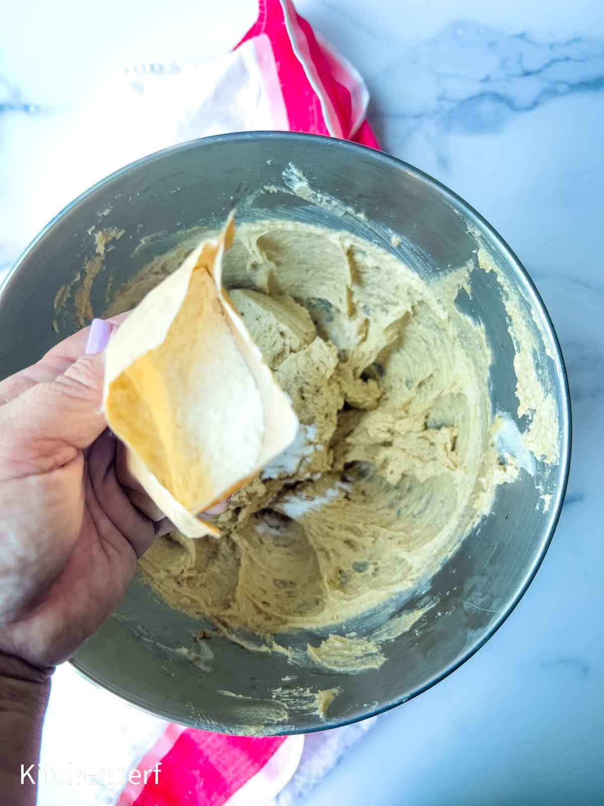 A hand adding flour from a bag into a mixing bowl with cookie dough batter on a marble countertop.