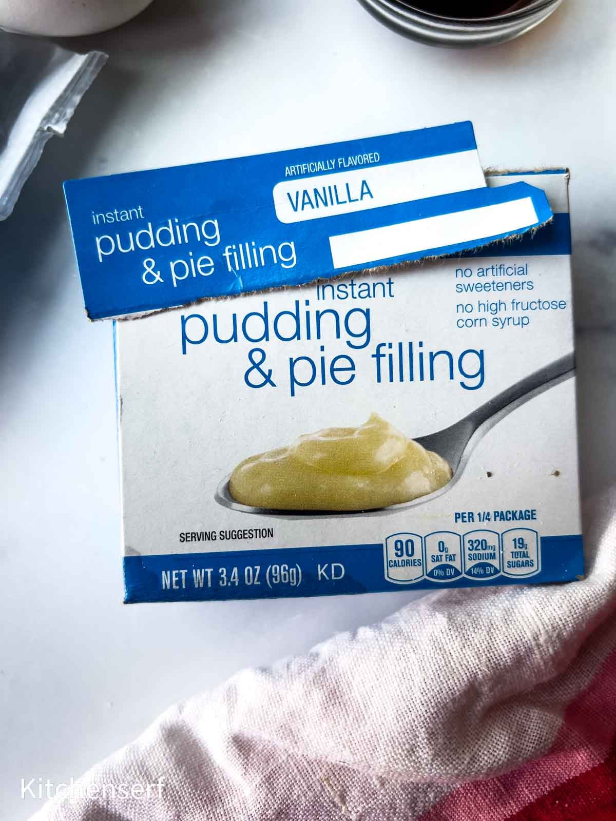 A box of instant vanilla pudding & pie filling on a white surface with a red-striped cloth beside it.