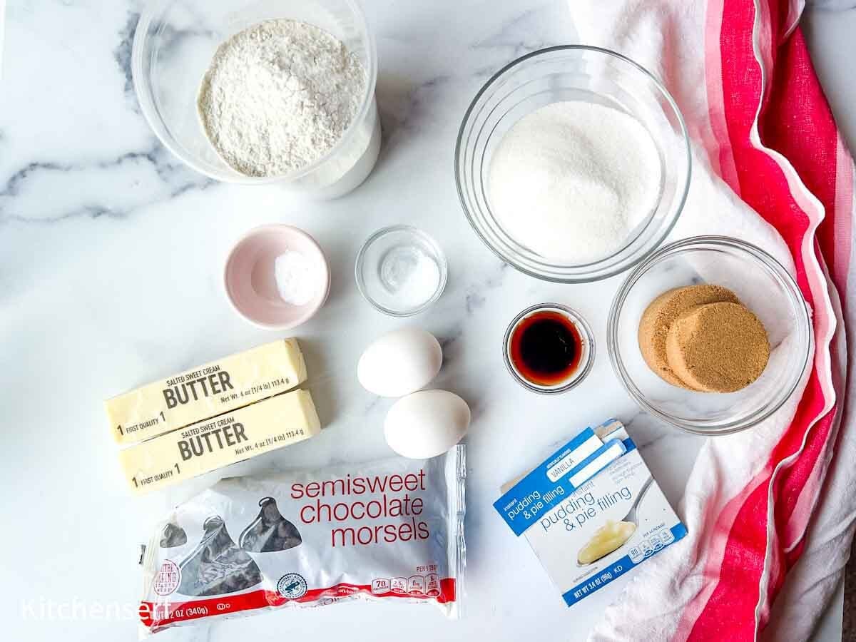 Baking ingredients on a counter: flour, sugar, brown sugar, butter, eggs, vanilla, chocolate chips, and pudding mix.
