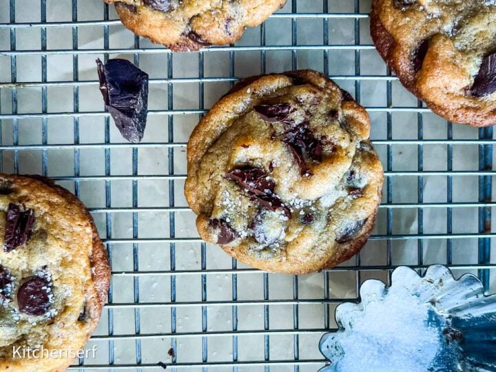 Chocolate chip cookies with vanilla pudding and flaky salt cool on a rack, surrounded by a bowl of salt and chocolate chunks.