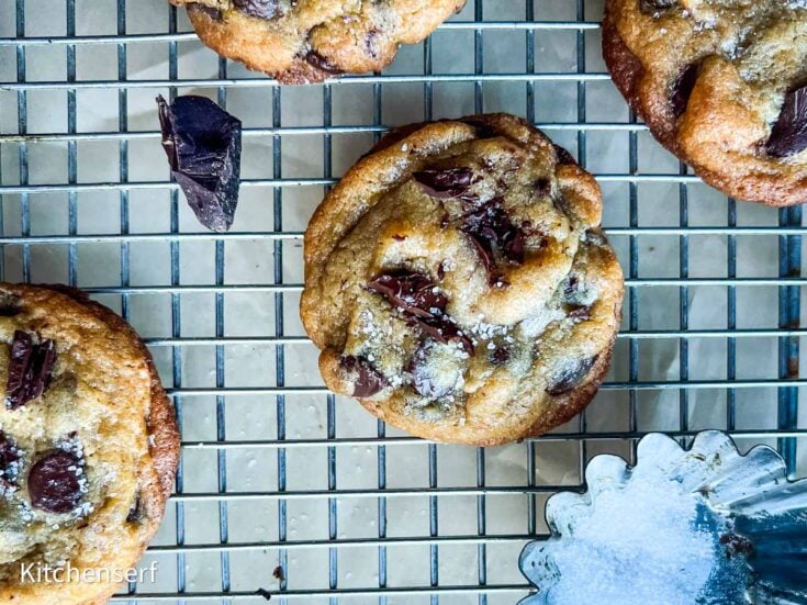 Chocolate chip cookies with vanilla pudding and flaky salt cool on a rack, surrounded by a bowl of salt and chocolate chunks.