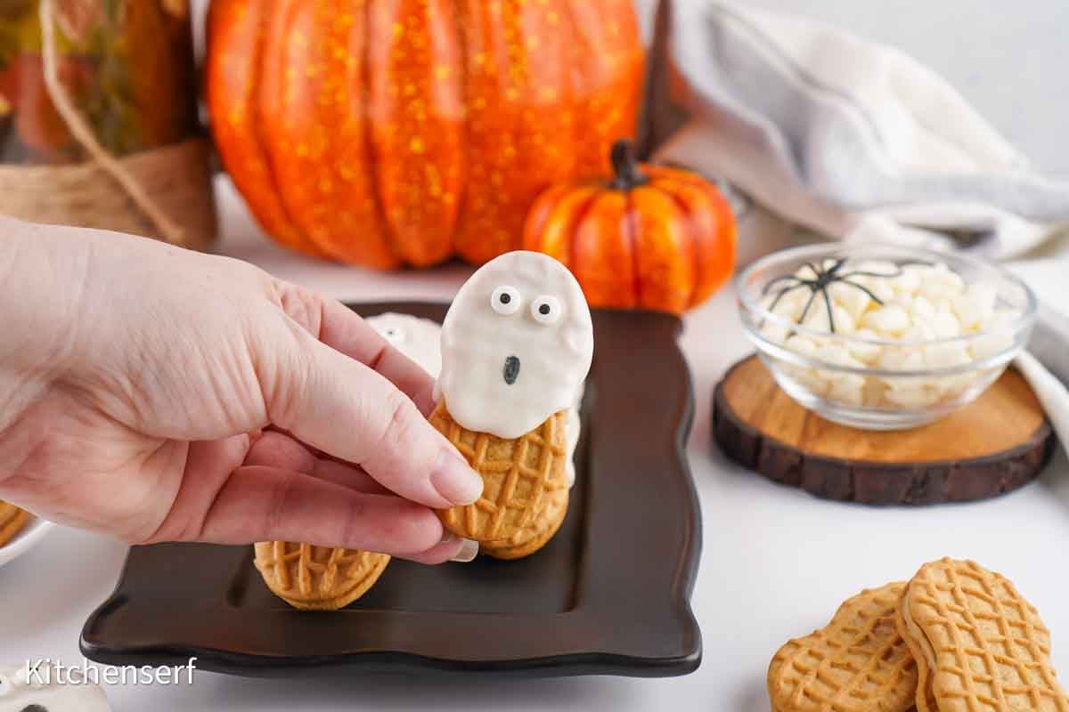 A hand holds a ghost-shaped cookie made from a peanut butter sandwich cookie dipped in white chocolate.