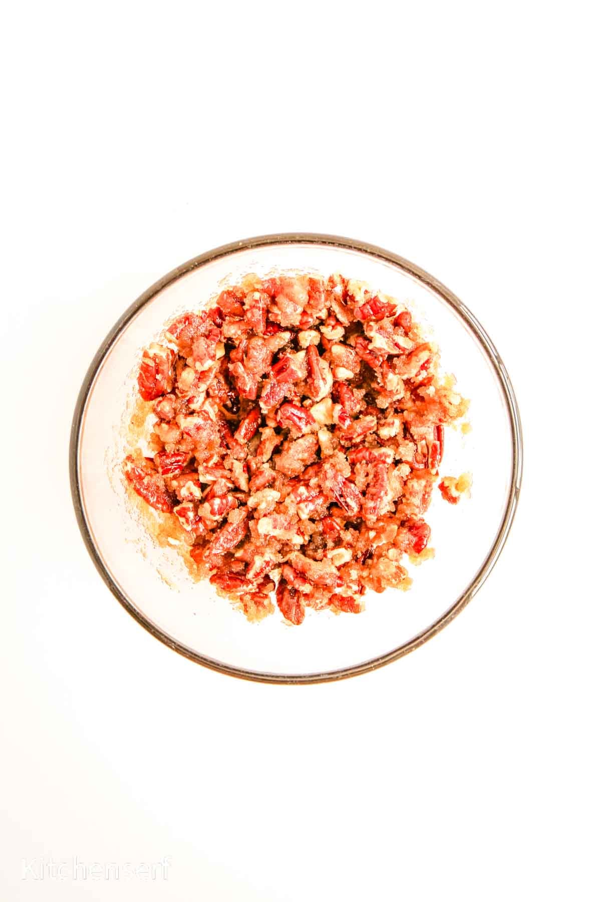 A glass bowl filled with chopped candied pecans, perfect for topping sits on a white background.