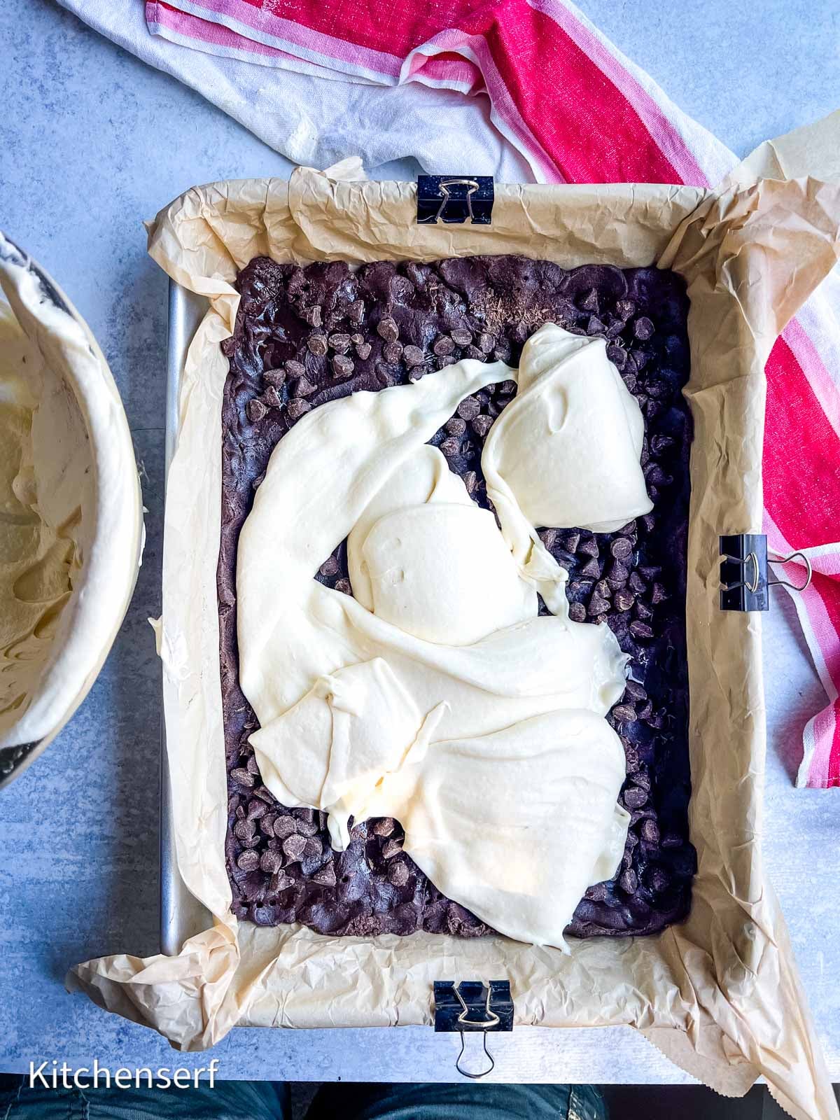 Brownie batter topped with chocolate chips and dollops of cream cheese in a parchment-lined baking pan.