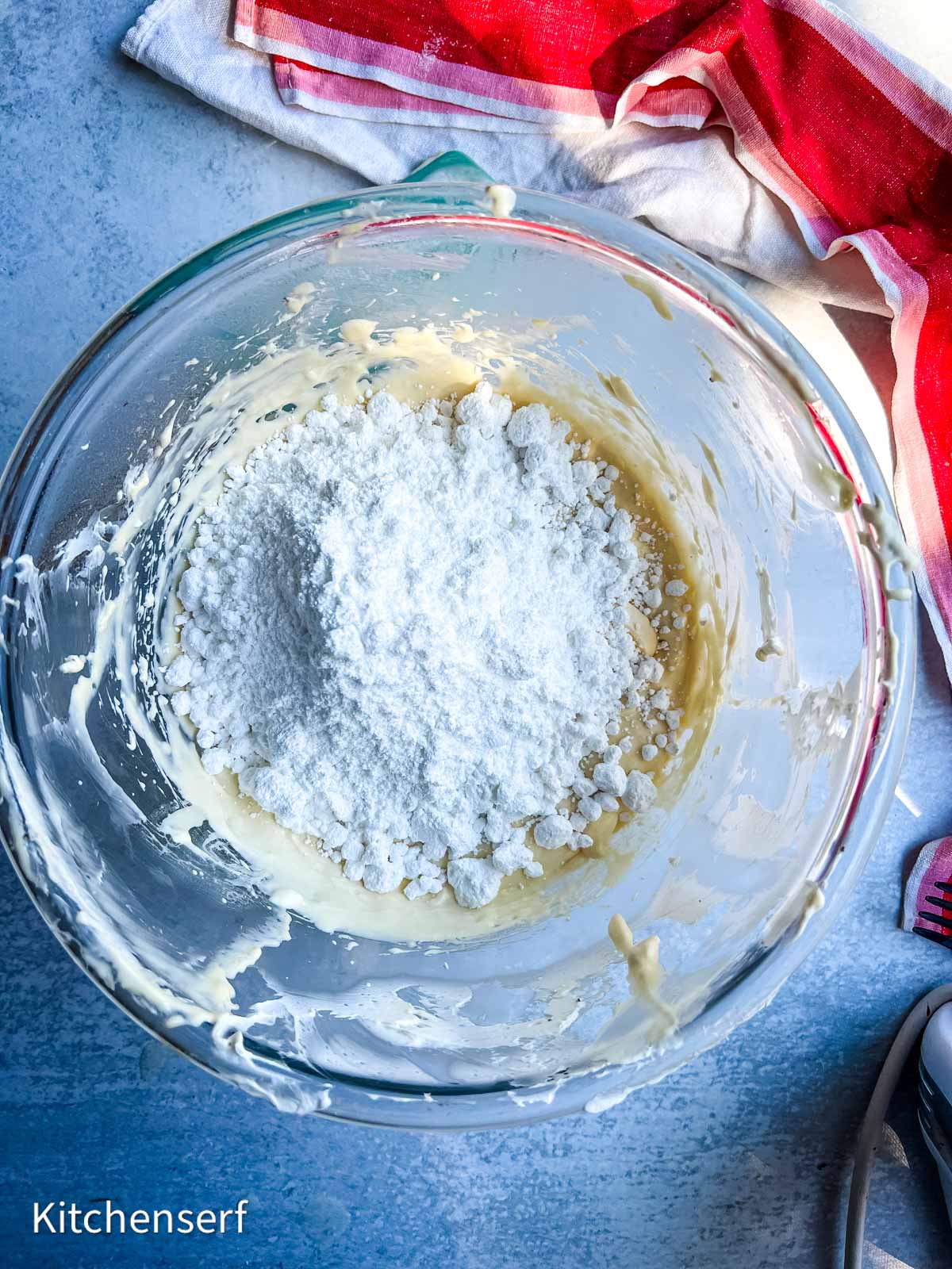A glass bowl with powdered sugar and batter being mixed, on a blue surface with a red and white towel nearby.
