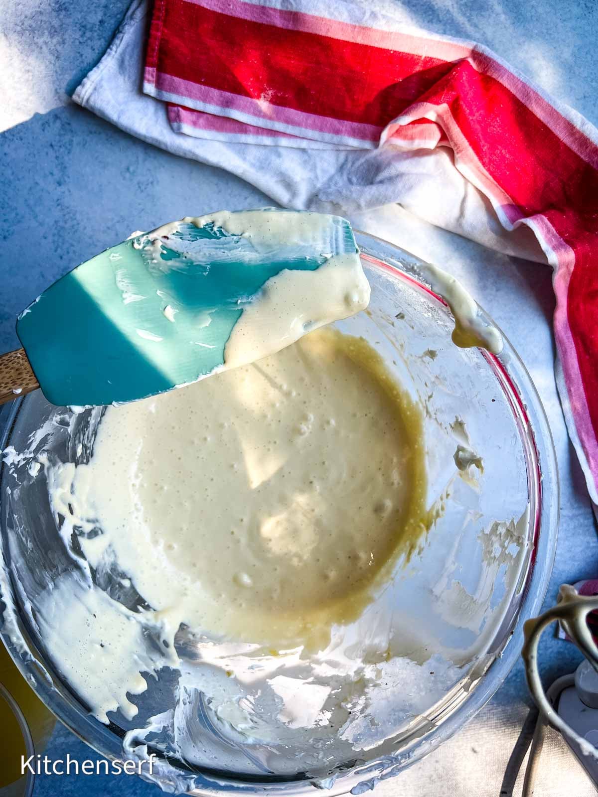 Glass bowl of cake batter with a blue spatula, red and white towel in background on a blue surface.