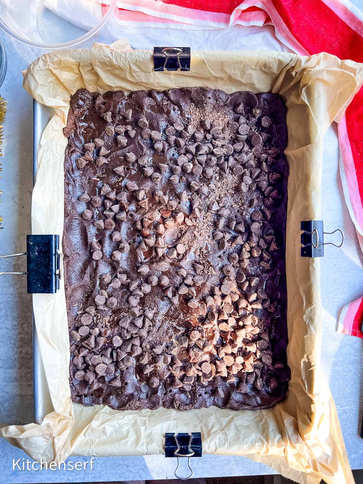 A pan lined with parchment paper holds chocolate brownies topped with chocolate chips.
