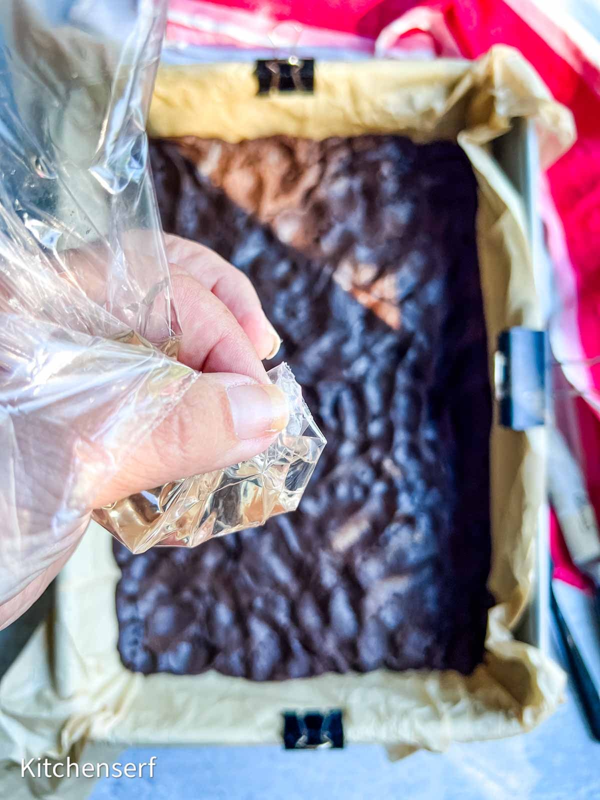 Hand pressing brownie dough in a parchment-lined pan using a plastic bag for even spreading.
