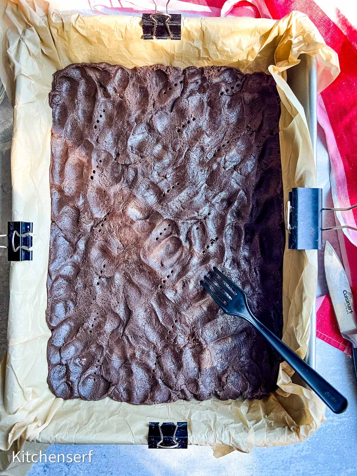 A baked brownie slab in a parchment-lined pan with a black fork resting on top.