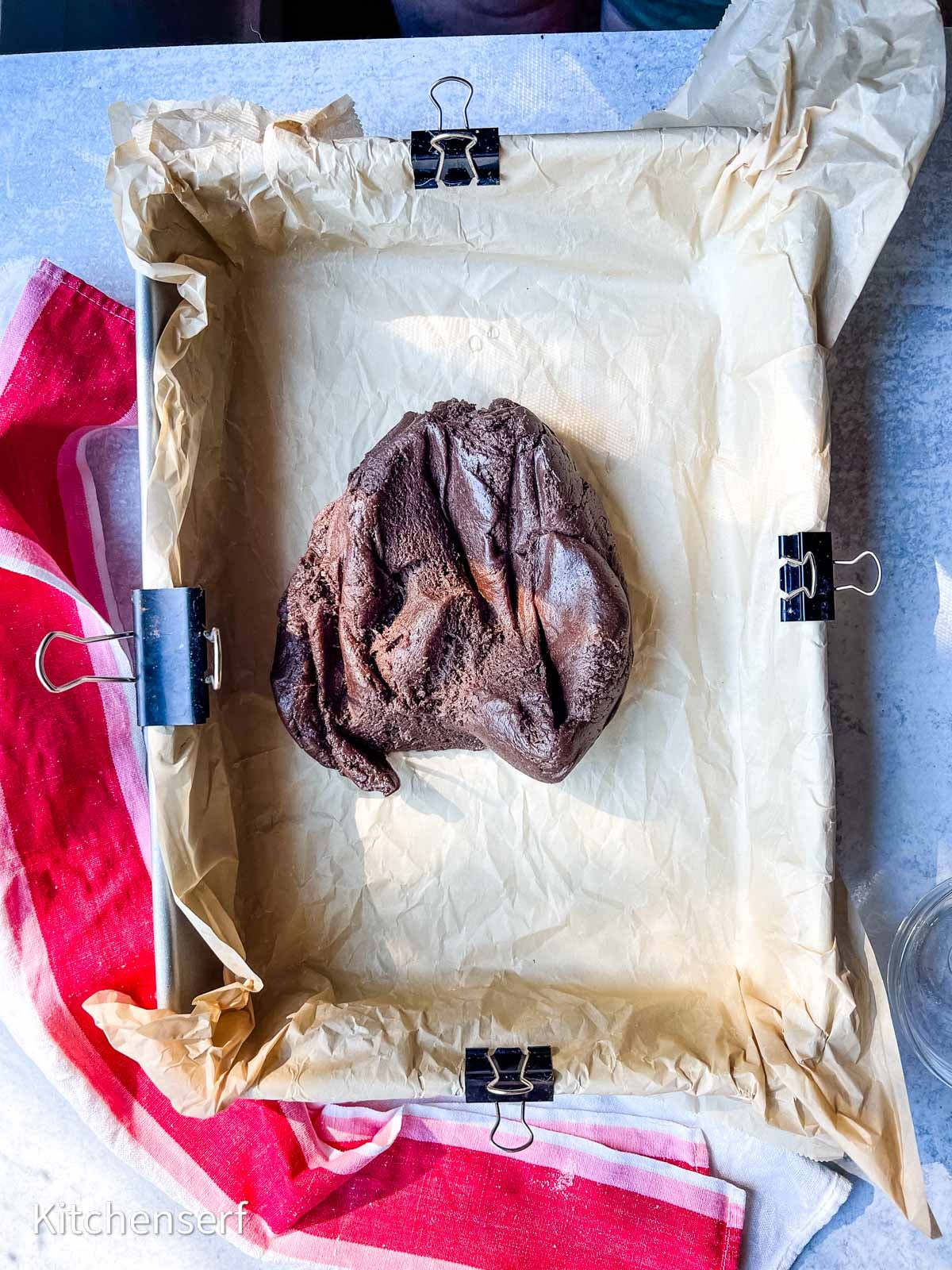 A ball of chocolate dough sits on parchment paper in a square baking pan with binder clips.