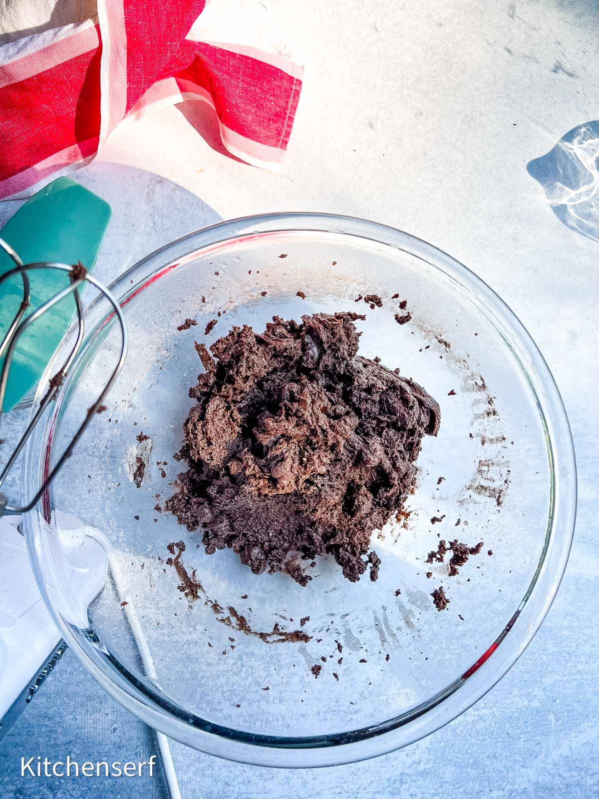 A glass bowl with chocolate cookie dough, a mixer, and a red-striped towel on a white surface.