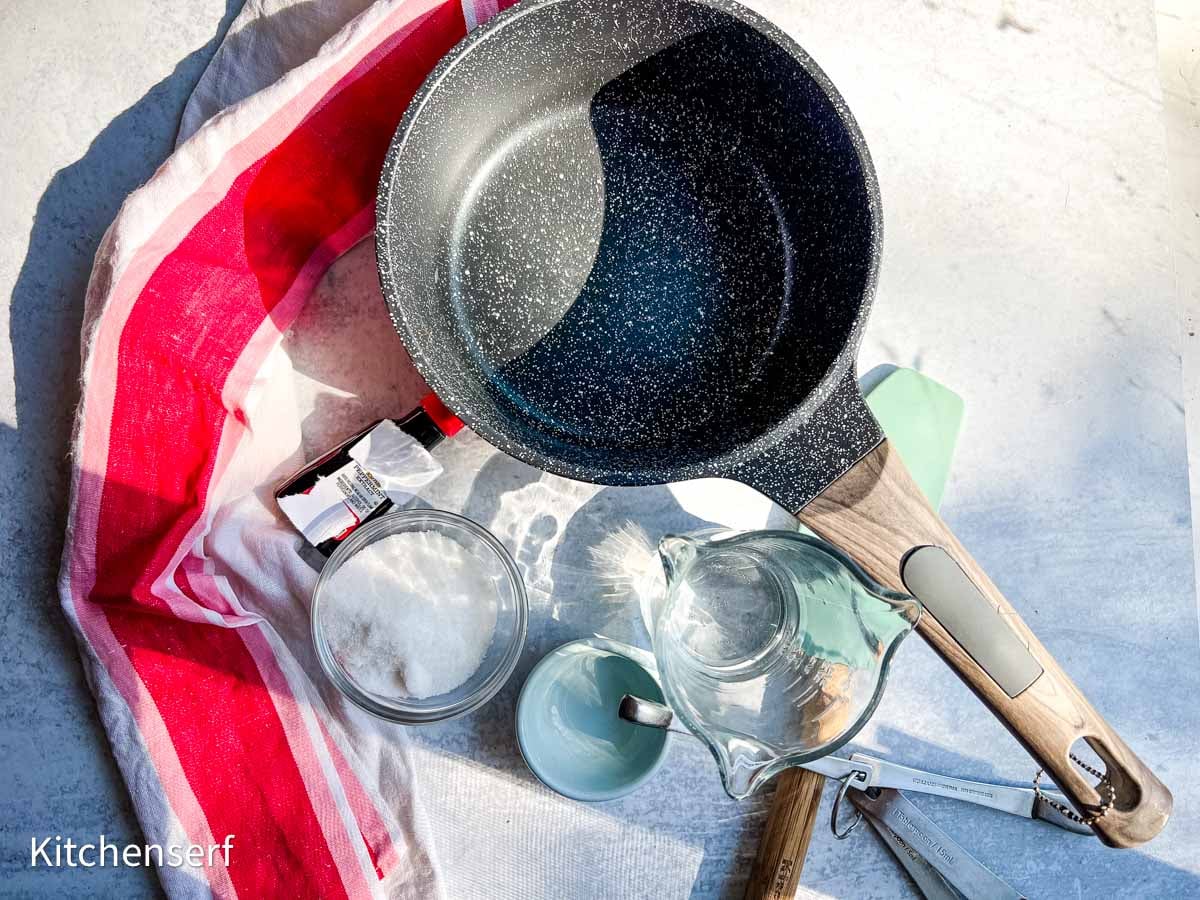 Nonstick pan, sugar, water, utensils, and a red-striped towel on a white surface in bright light.