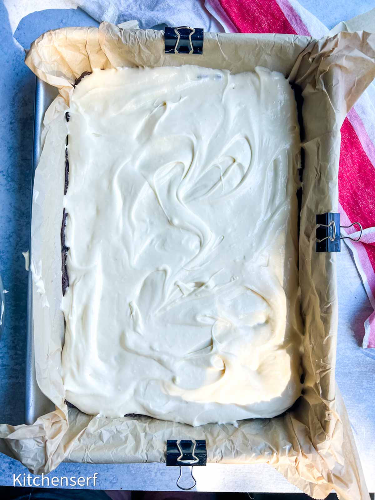 A rectangular cake with gooey butter topping  in a parchment-lined pan, secured with metal binder clips.
