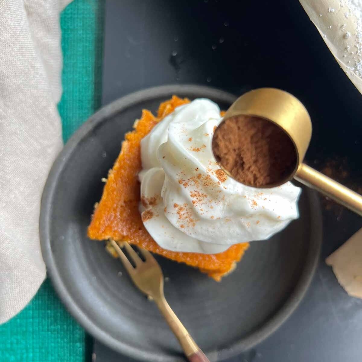 slice of pumpkin cake topped with whipped cream on a black plate with a teaspoon of pumpkin pie spice overhead.