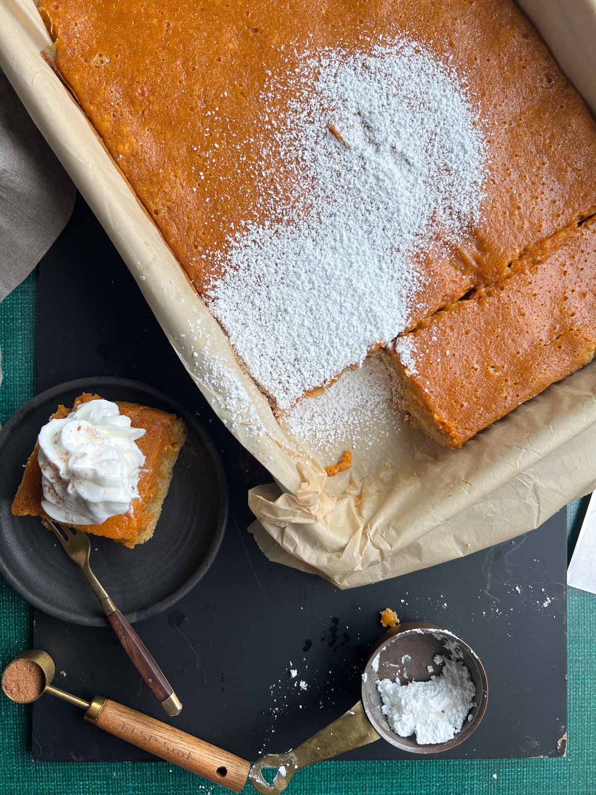 slice of pumpkin gooey butter cake next to a parchment lined pan of pumpkin gooey butter cake with powdered sugar over the top.
