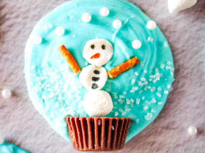 Blue frosted cookie topped with a snowman made of marshmallows, pretzel arms, and candy decorations.