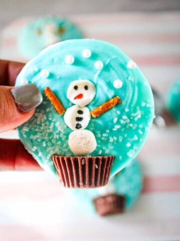 A hand holds a blue frosted cookie decorated with a snowman made of candy and pretzels.