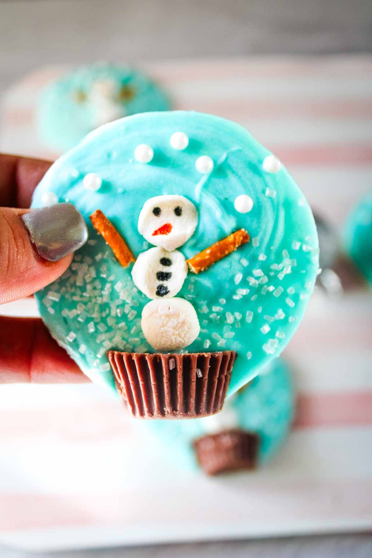A hand holds a blue frosted cookie decorated with a snowman made of candy and pretzels.