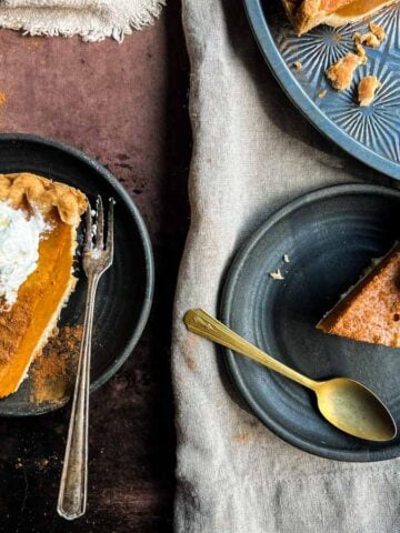 Two slices of pumpkin pie with whipped cream on black plates, with a fork and spoon beside each slice.