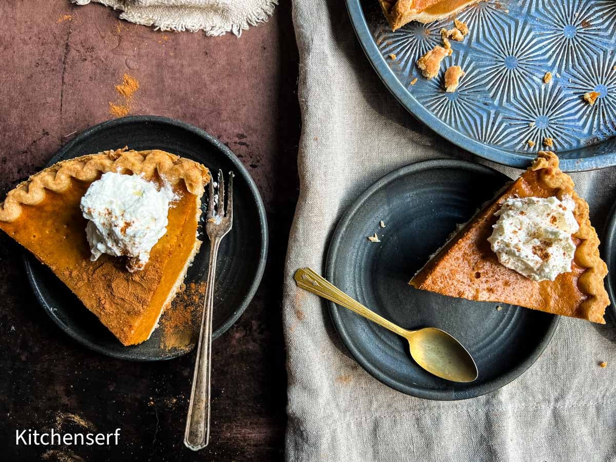 Two slices of pumpkin pie with whipped cream on black plates, with a fork and spoon beside each slice.
